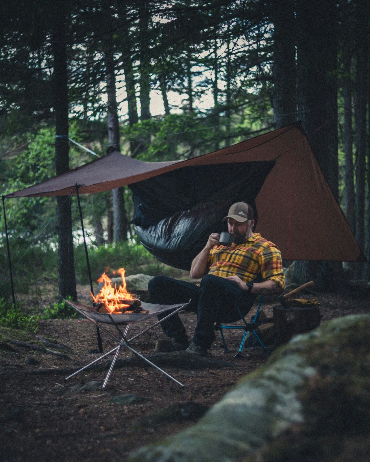 En man dricker kaffe vid elden framför sin hammock och tarp i skogen