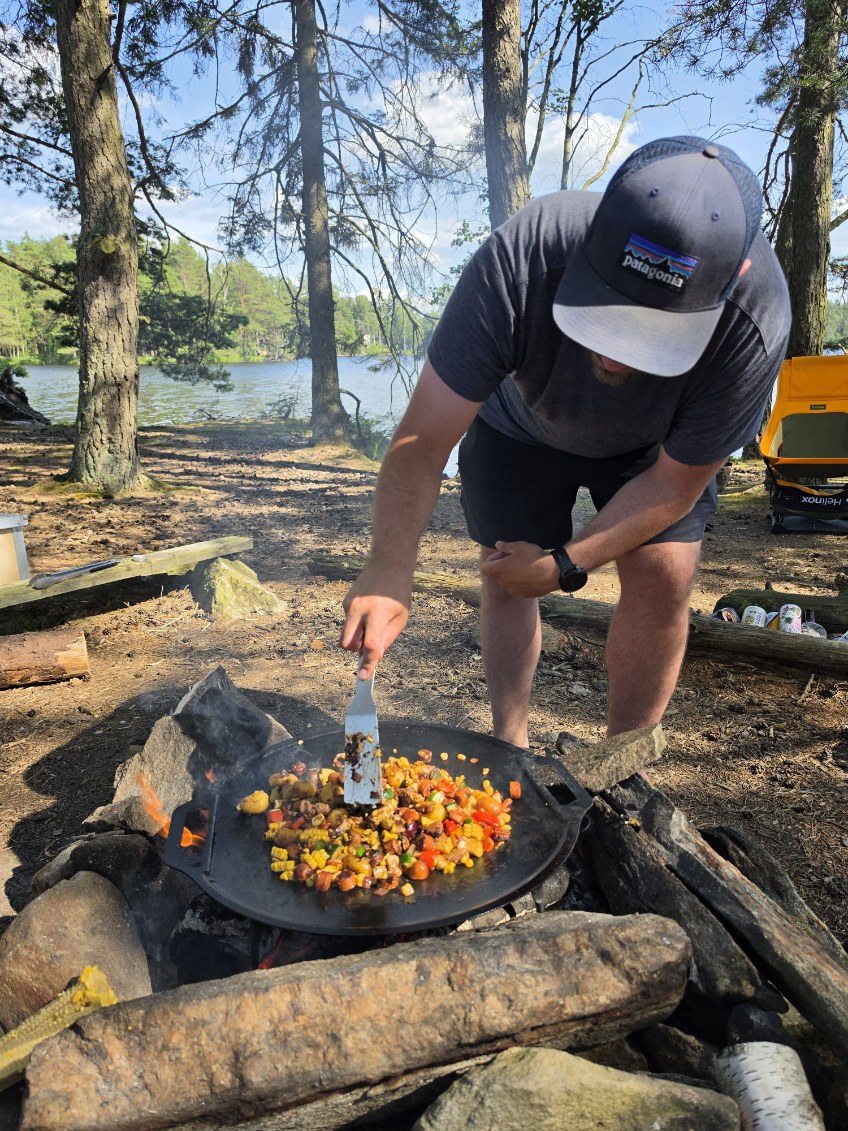 En man lagar middag på en stor stekhäll över elden, med sjön och tallskogen bakom